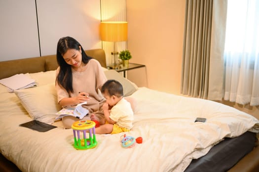 Asian mother working remotely on a laptop, cute baby boy playing toys on the bed next to her mother.