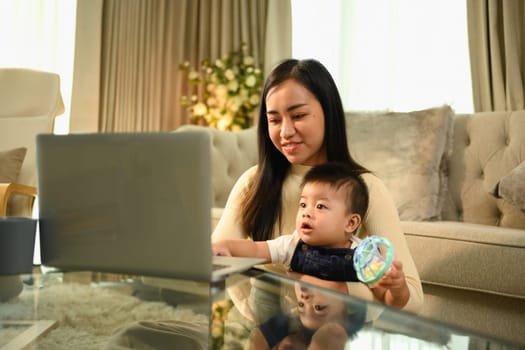 Young asian mother holding her baby boy on hands while working with laptop at home.