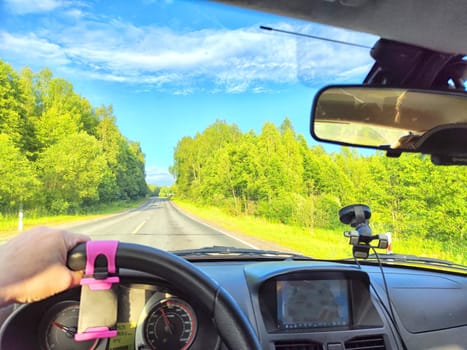 Car salon, windshield, hand of woman on steering wheel and landscape. View from seat of driver on nature with Road, trees, blue sky at a sunny day. Single trip of female traveler. Partial focus