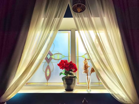 Curtains frame a sunlit window with vibrant red flowers on sill. Bright Morning Sunlight Through Window With Red Flowers. Background. Partial focus