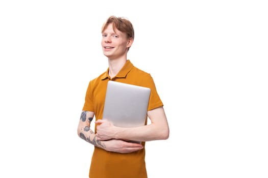 young man with red hair in an orange t-shirt holding a laptop.