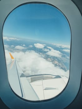 vertical photo of an airplane window, view of the wing, blue sky in the clouds and mountains.
