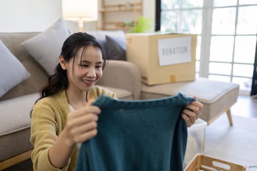 Young Asian women sit in living room sorting clothes for donation in a donation box second hand clothes.