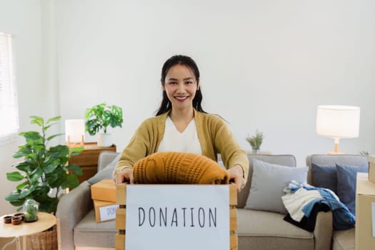 Young Asian women sit in living room sorting clothes for donation in a donation box second hand clothes.