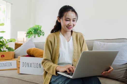 Asian young woman using laptop for donate box with second hand clothes.