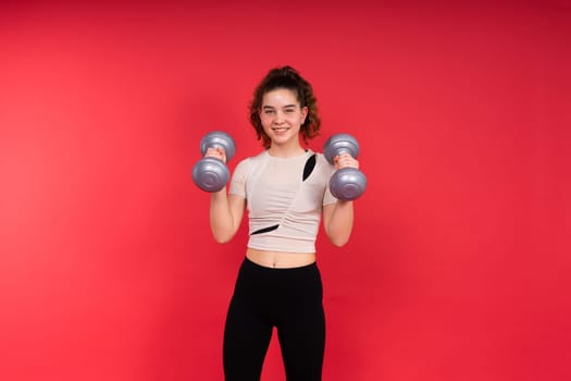 Teenage sportive girl exercises with dumbbells to develop muscles isolated on a red background.