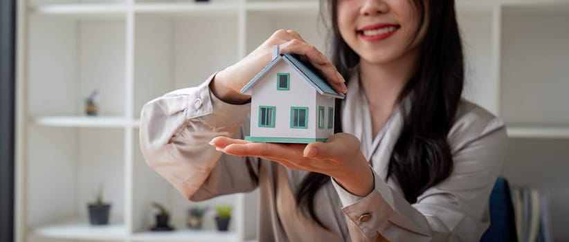 Smiling real estate agent woman hands protected miniature house on wooden desk. Property insurance and reputable finance calm concept.