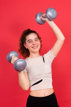 Teenage sportive girl exercises with dumbbells to develop muscles isolated on a red background.