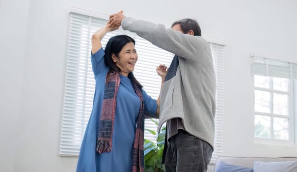 Elderly Asian couple in their 60s dancing in the living room together, enjoying music and smiling in love..