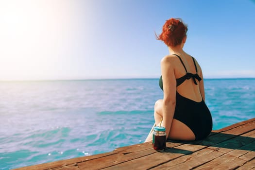 Attractive girl enjoys sea view on vacation. Red-haired woman in swimsuit is sitting on edge of a wooden pier with a bottle of refreshing lemonade next to her. Tourism and travel. Sunlight at sky.