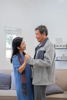 Elderly Asian couple in their 60s dancing in the living room together, enjoying music and smiling in love..