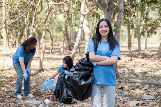 Portrait of a cute young woman holding a garbage bag with a group of Asian volunteers helping to collect garbage in plastic bags, cleaning up the area in the forest to preserve the natural ecosystem..