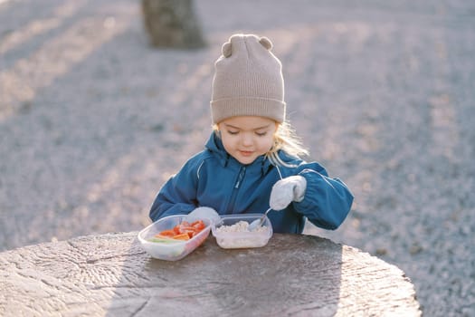 Little smiling girl scoops porridge with a spoon from a lunchbox while standing behind a tree stump. High quality photo