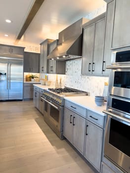 Denver, Colorado, USA-March 3, 2024-This modern kitchen showcases professional-grade stainless steel appliances, including a high-end gas range and double ovens, set against gray shaker cabinets and a marble backsplash. The warm wood flooring complements the cool tones of the cabinetry, creating an inviting culinary space.
