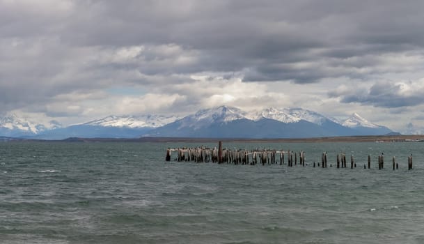 Wooden posts in choppy sea before snowy mountains.