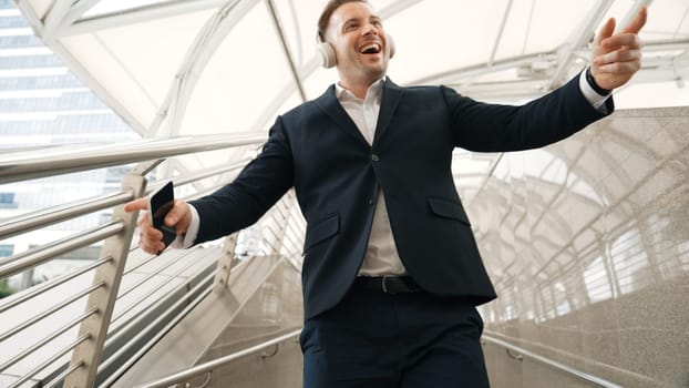 Business man dance to relaxing music from headphone at modern corridor. Cheerful project manager wear headset and moving gesture to celebrate successful song with lively mood in urban city. Urbane.