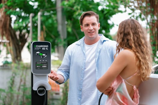 Happy and lovely couple with eco-friendly conscious recharging electric vehicle from EV charging station. EV car technology utilized as alternative transportation for future sustainability. Synchronos