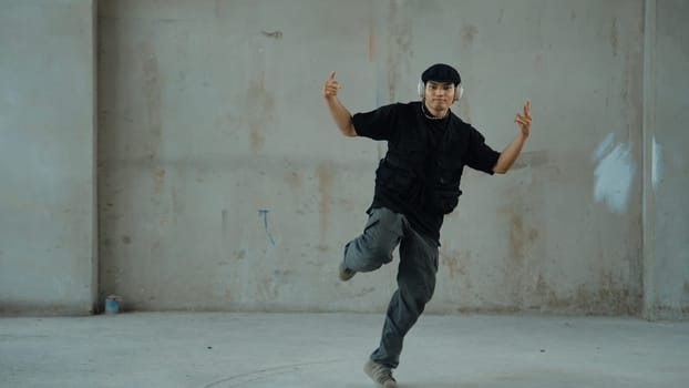 Handsome hipster practicing break dancing while listening music from headphone with cement background. Motion shot of street dancer perform robot dance. Outdoor sport 2024. Modern dance. Endeavor.