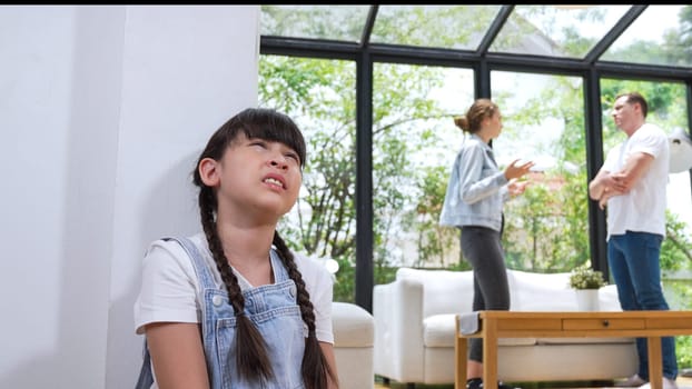 Stressed and unhappy young girl huddle in corner, cover her ears blocking sound of her parent arguing in background. Domestic violence at home and traumatic childhood develop to depression. Synchronos