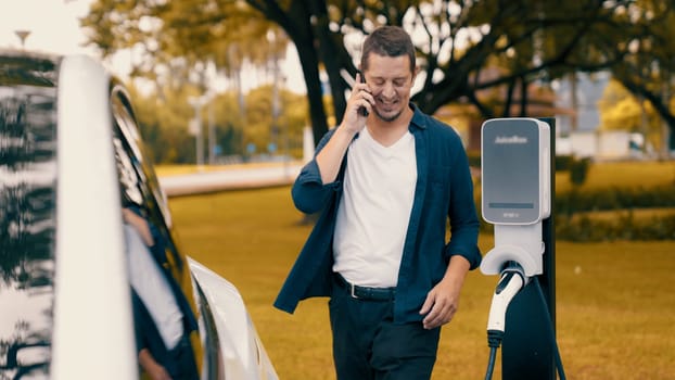 Man recharging battery for electric car during road trip travel EV car in autumanl season natural forest or national park. Eco friendly travel during vacation and holiday. Exalt