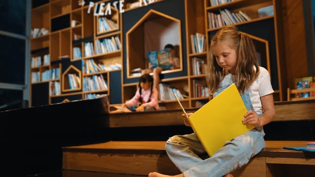 Young smart caucasian girl picking reading a book while sitting at library. Clever child learning, studying, open a books at library. Attractive kid turning page with blurring background. Erudition.