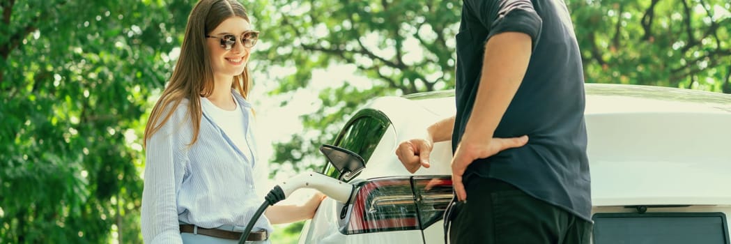 Focused EV car recharging electricity for battery on blurred background of lovey couple ruing their road trip travel by eco friendly electric car in national park and greenery forest on holiday. Exalt