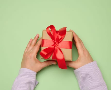 Woman's hand holding a gift box wrapped in a red silk ribbon on a green background, top view