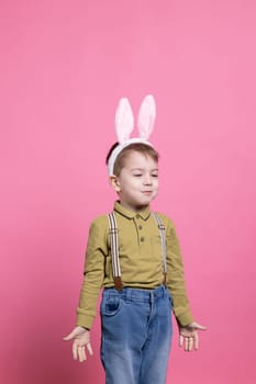 Small boy wearing bunny ears in front of the camera, feeling excited about easter holiday festivity and receiving gifts. Innocent young child smiling and feeling happy against pink background.