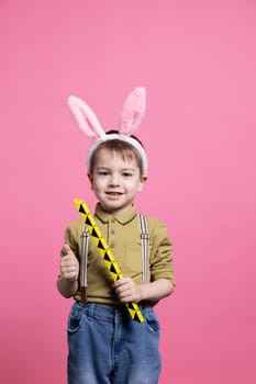 Young cute toddler playing around with a toy in front of camera, feeling happy while he wears fluffy bunny ears. Joyful small child posing against pink background, innocent kid.