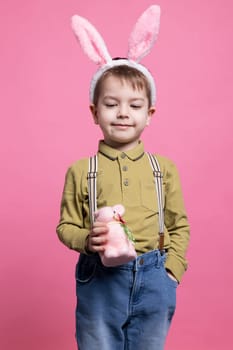 Young small child smiling in studio and holding a cute pink rabbit toy, celebrating easter holiday with handmade painted decoration. Joyful little boy wearing bunny ears and poses on camera.