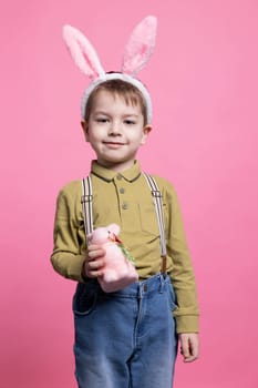 Joyful little boy holding a fluffy stuffed rabbit in front of on camera, wearing bunny ears and feeling excited about easter celebration. Small kid smiling and being happy against pink backdrop.
