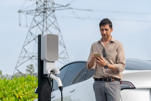 Man pay for electricity with smartphone while recharge EV car battery at charging station connected to power grid tower electrical as electrical industry for eco friendly car utilization.Expedient