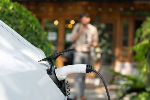 Focused EV electric car recharging at outdoor coffee cafe in springtime garden with blur background of eco friendly man, green city sustainability and environmental friendly EV vehicle. Expedient