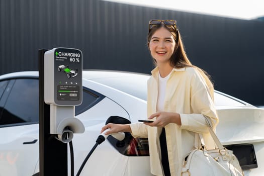 Young woman holding shopping bag and use smartphone to pay for electricity for recharging EV car battery from charging station at city mall parking lot. Modern woman go shopping by eco car. Expedient