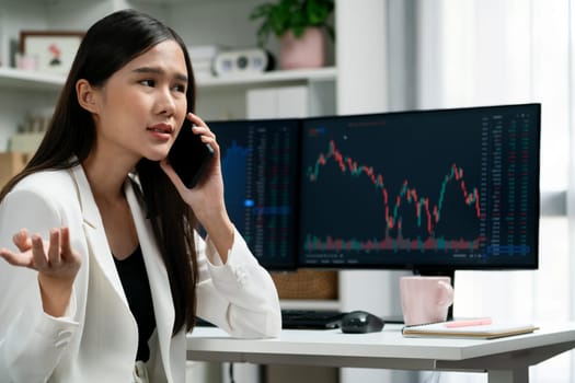 Stressful young beautiful Asian businesswoman calling to broker on smartphone, analyze possible risk to profit or loss in dynamic exchange stock market graph on pc screen at modern office. Stratagem.