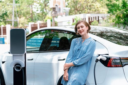 Beautiful young woman recharging her electric car from home EV charging station using alternative energy with net zero emission. Modern young girl charging her vehicle before vacation travel.Perpetual