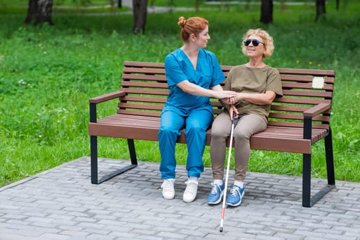 A nurse and an elderly blind woman are sitting on a bench in the park
