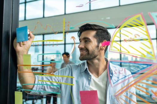 Portrait of young handsome businessman writing marketing idea by using colorful marker on glass wall with graph and mind map. Manager sharing idea or financial strategy. Business plan. Tracery.