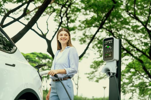 Young woman recharging battery for electric car during road trip travel EV car in natural forest or national park. Eco friendly travel during vacation and holiday. Exalt