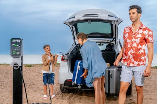 Family vacation trip traveling by the beach with electric car, lovely family taking luggage out while charging EV car battery with clean energy. Alternative family travel by eco-friendly car.Perpetual