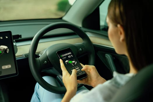 Young woman inside the vehicle, checking EV vehicle's application on battery recharging electricity status display on smartphone screen in modern EV car on her road trip journey. Exalt