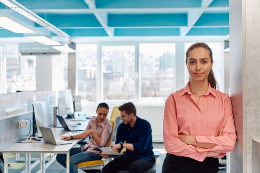 Portrait of young smiling business woman in creative open space coworking startup office. Successful businesswoman standing in office with copyspace. Coworkers working in background.