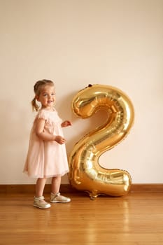 Little happy girl in a festive dress stands near an inflatable golden number in the room. High quality photo