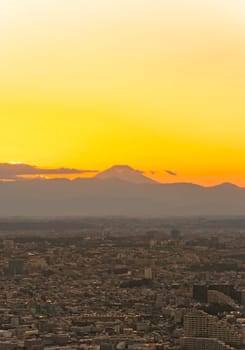 Magnificent sunset at golden hour overlooking an urban cityscape of the megalopolis of Tokyo whose tiny buildings as far as the eye can see contrast with the large cone of Fuji mountain on the horizon