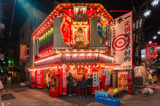 osaka, japan - dec 04 2023: Illuminated shooting game attraction store in the Tsutenkaku Hondori Shopping Street of Shinsekai district adorned with a golden statue of Biliken and yōkai demon at night.