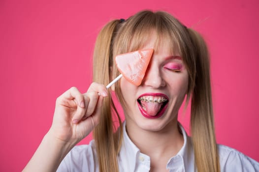 Portrait of a young woman with braces and bright makeup eating a lollipop on a pink background