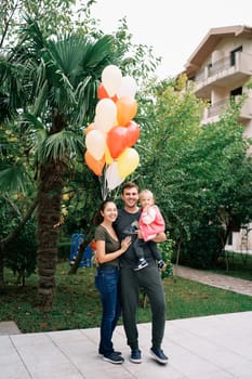 Smiling mom hugging dad with a little girl in his arms in the garden near colorful balloons. High quality photo