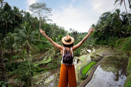 Back view of happy woman with arms up enjoying vacation in Tegalalang rice terrace, Bali. Female enjoying freedom in nature. Trip, vacation, mental health and healthy lifestyle concept.