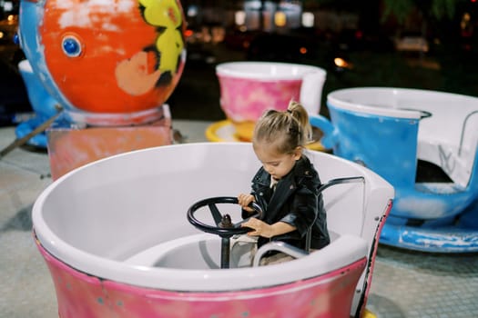 Little girl sits at the wheel of a cup-shaped carousel and looks at her feet. High quality photo