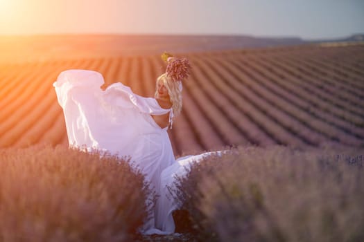 Blonde woman poses in lavender field at sunset. Happy woman in white dress holds lavender bouquet. Aromatherapy concept, lavender oil, photo session in lavender.